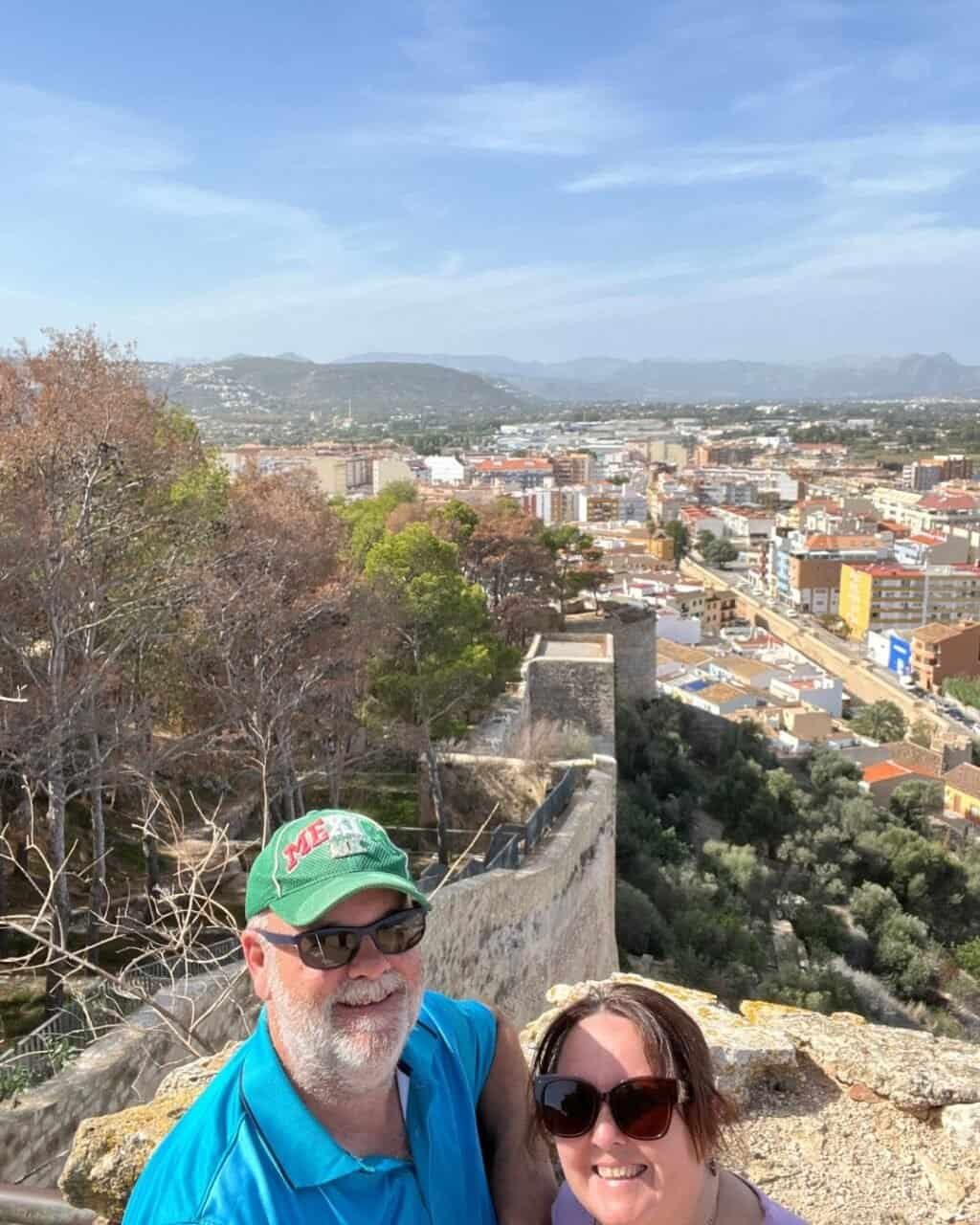 a couple standing at the top of the denia castle walls with the city below